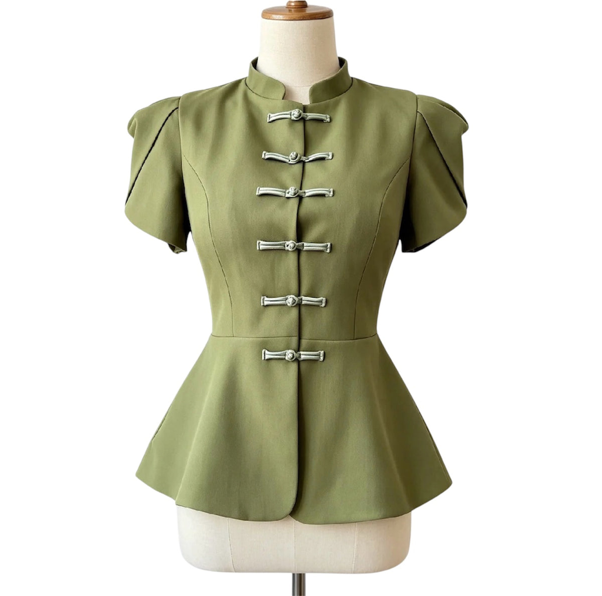 An olive green short-sleeve peplum top with silver metal toggle closures, displayed on a cream-colored dress form against a white background.