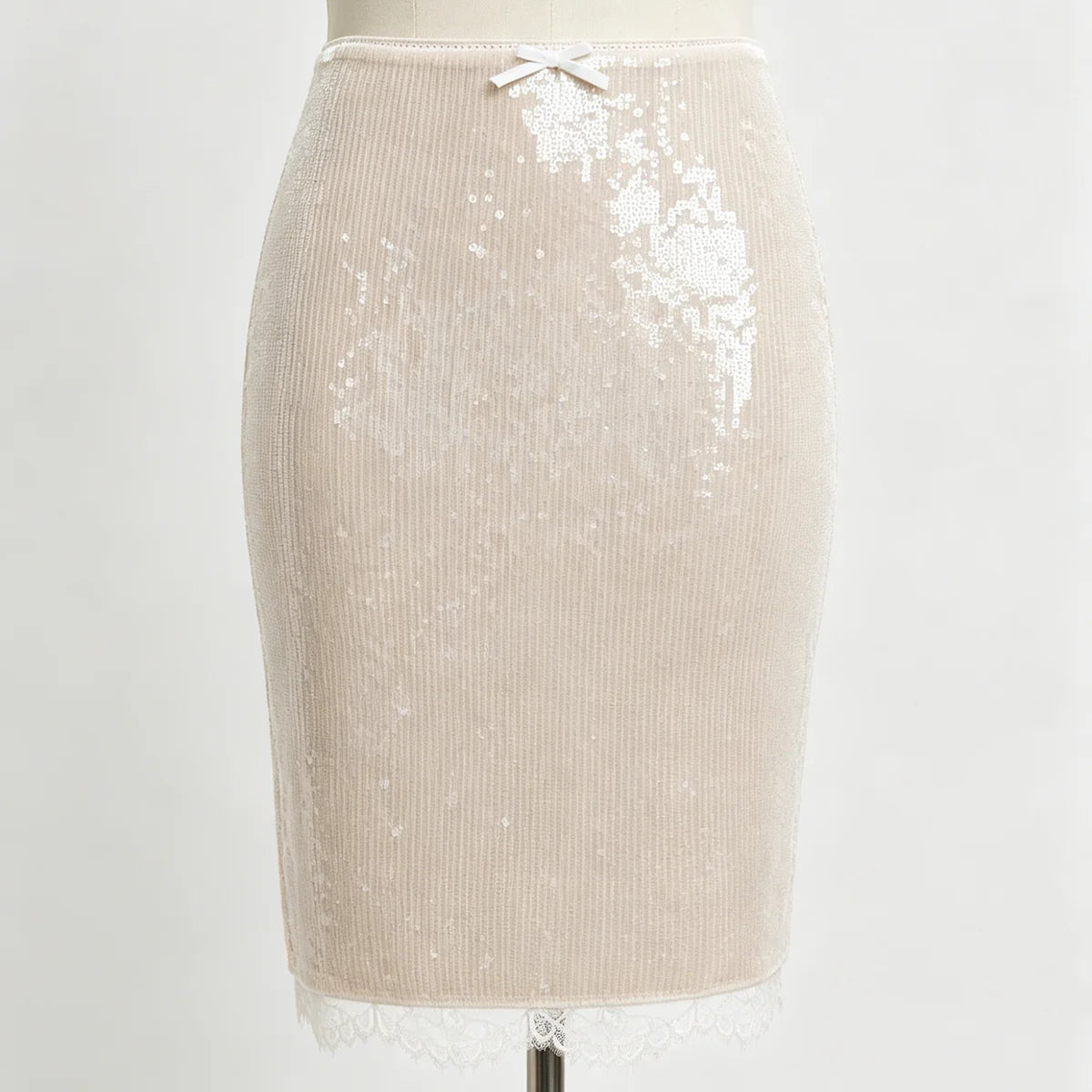 A beige ribbed knit pencil skirt with white sequins, a small white bow at the waistband, and a white lace hem, displayed on a dress form against a white background.