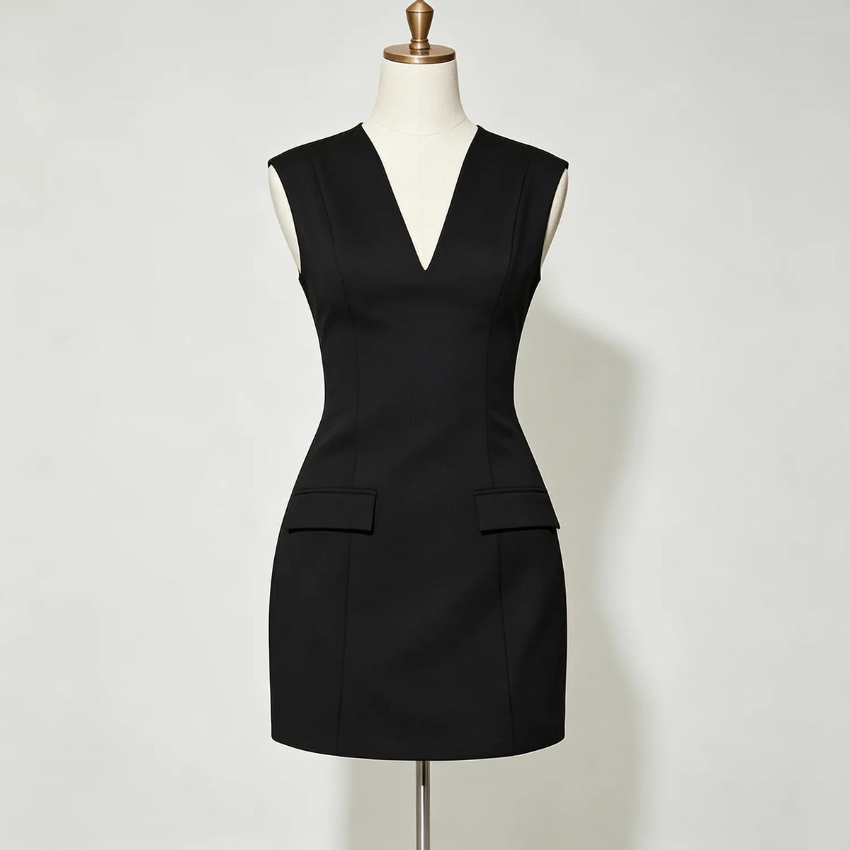 A sleeveless black mini dress with a deep V-neckline and two flap pockets at the waist, displayed on a cream-colored dress form mannequin against a light gray background.
